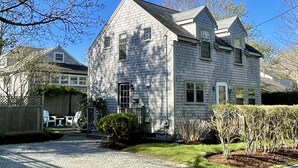 A two-story house with a stone foundation, a wooden deck, and a well-maintained lawn. - Bright, In-Town Nantucket Cottage + Yard - Walk to Main St. (Nantucket)