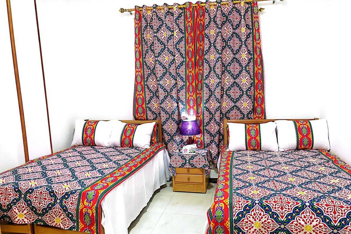 A room with two single beds, a wooden nightstand, a purple lamp, and patterned curtains.