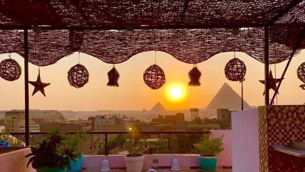 A rooftop terrace with a view of the pyramids at sunset, featuring a round table, colorful cushions, and potted plants. - Gohar Guest House (Giza)