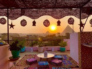 A rooftop terrace with a view of the pyramids at sunset, featuring a round table, colorful cushions, and potted plants. - Gohar Guest House (Giza)