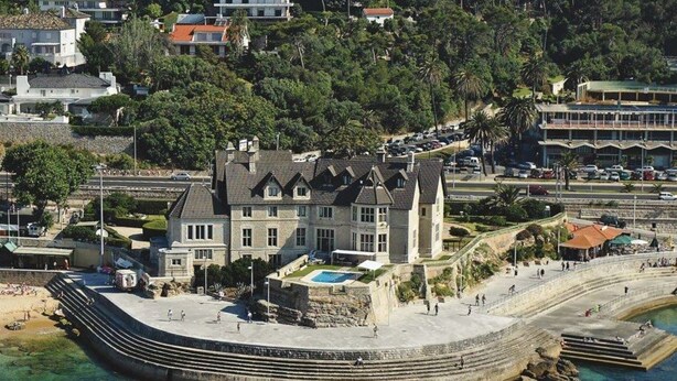 Un edificio frente al mar con una amplia piscina, escalones de piedra que bajan hacia el agua y vistas al océano.