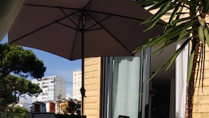Una piscina con suelo de baldosas, un área de patio con sillas y una sombrilla, y vistas de edificios al fondo.