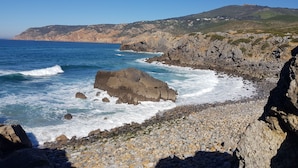 Una costa rocosa con olas rompiendo en la orilla, cielo despejado y una colina distante con edificios.