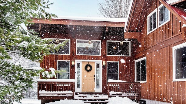 A wooden cabin with a front porch, a door with a wreath, and a snow-covered landscape.