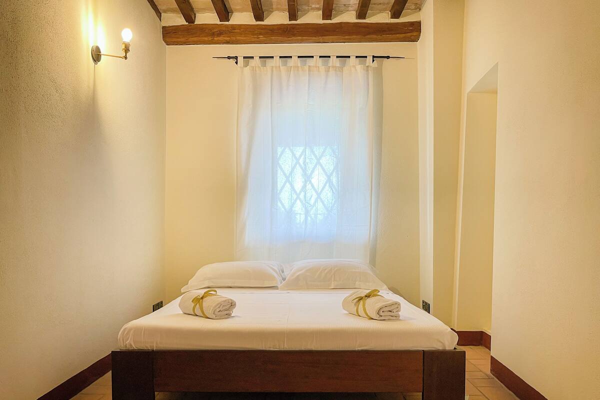 Une chambre d’hôtel avec un lit en bois, du linge de lit blanc, une fenêtre ornée d’un motif en treillis et un luminaire fixé au mur.