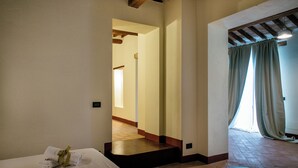 A hotel room with a bed, a small table with a towel, a wooden beam ceiling, and a view through a window with curtains. - Villaluce Wine Agriturismo (Castiglione del Lago)