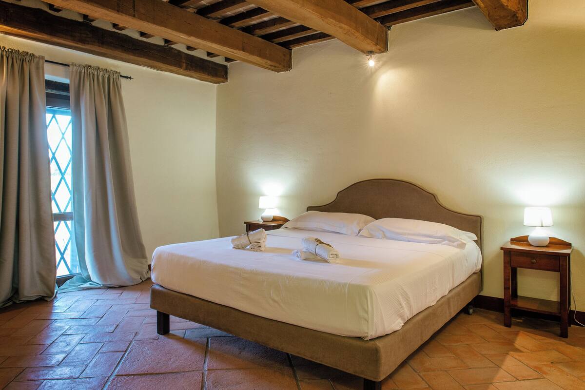 Un lit bien fait, avec du linge de lit blanc et deux oreillers, une table de chevet en bois avec une lampe, et une fenêtre avec des rideaux.