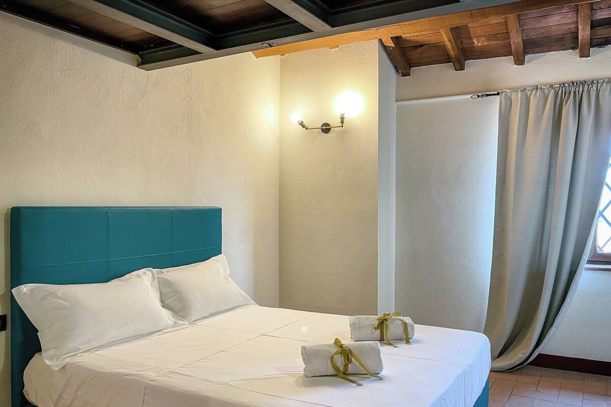 Un lit bien fait, avec du linge de lit blanc et une tête de lit turquoise, deux serviettes ornées de rubans et un luminaire fixé au mur.