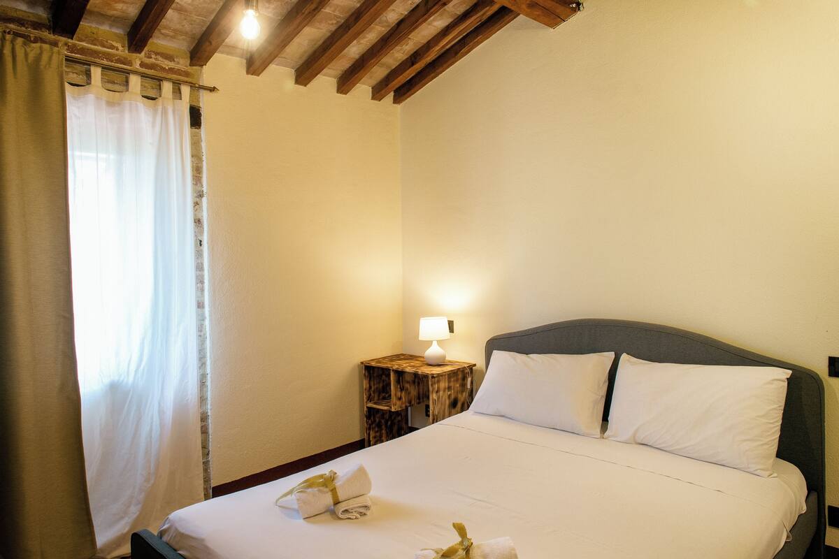 Un lit bien fait, avec du linge de lit blanc et des oreillers, une table de chevet en bois avec une lampe, et une fenêtre avec des rideaux.