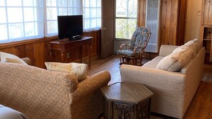 Uma sala de estar acolhedora com paredes e pisos de madeira, uma televisão sobre um suporte de madeira, dois sofás, uma poltrona estampada e uma pequena mesa de madeira.