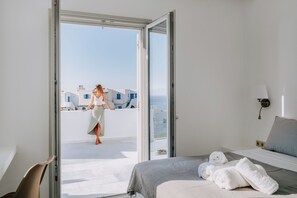 A hotel room with a bed, a chair, a view of the sea, and a balcony with a person. - SUNSENSES VILL ARIADNE WITH PRIVATE POOL (Paros)