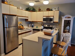 A compact kitchen with a stainless steel refrigerator, microwave, and stove, a small island with a basket, and a white chair.