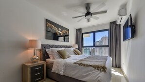 Ein modernes Schlafzimmer mit Bett, Nachttischen, Deckenventilator, großem Fenster und an der Wand montiertem Fernseher.
