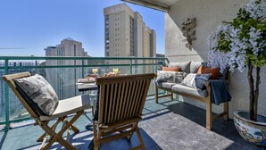 Ein Balkon mit Glasgeländer, einem Holzstuhl, einer Couch, einem Tisch mit Getränken und einem Blumentopf.