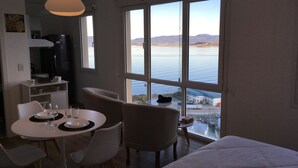 Ein Hotelzimmer mit großem Fenster, das Blick auf das Meer und sanfte Hügel bietet. Im Inneren befinden sich ein Esstisch mit Stühlen, ein Bett und eine Küchenzeile.