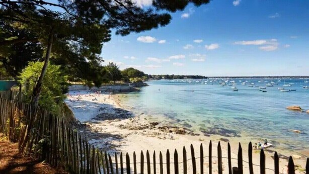A beach with clear water, a wooden fence, and boats in the distance. - APARTMENT IN MORBIHAN - DIRECT ACCESS TO THE PORT AND BEACH ON FOOT (Arzon)
