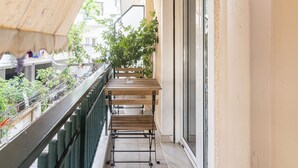 Un balcón estrecho con un banco de madera, barandilla metálica y vistas a una calle con áreas verdes y edificios.