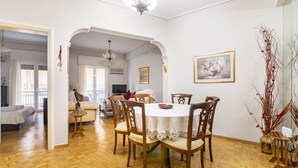 Un comedor con una mesa redonda, sillas de madera y un mantel blanco. A la derecha, un jarrón con ramas secas.