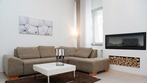 A modern living room with a sectional sofa, a white coffee table, a fireplace with logs, a mounted TV, and a wall art piece. - Bright, chic apartment in the heart of Düsseldorf near the main train station (Düsseldorf)