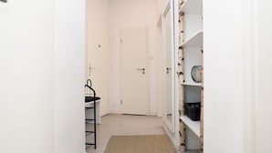 A hallway with a white door, a black chair, and a wooden shelf with baskets. - Bright, chic apartment in the heart of Düsseldorf near the main train station (Düsseldorf)