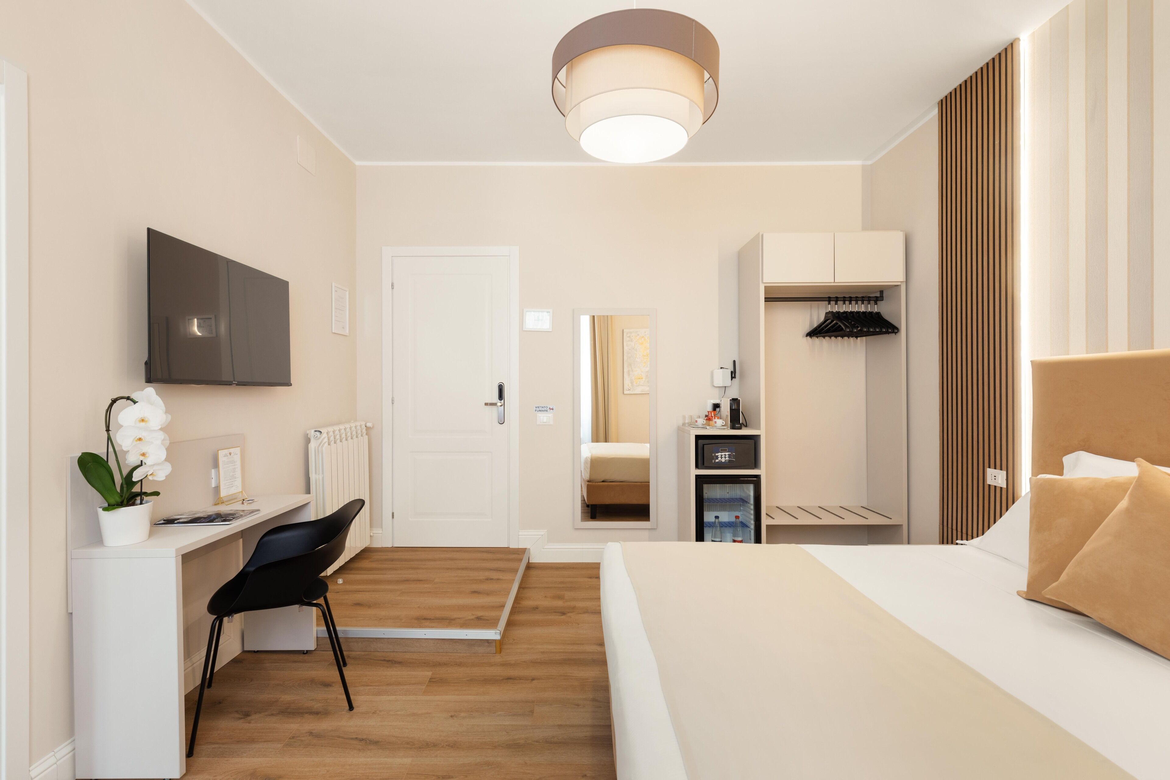 A hotel room with a bed, a desk with a chair, a TV, a radiator, a wardrobe, and a mirror.