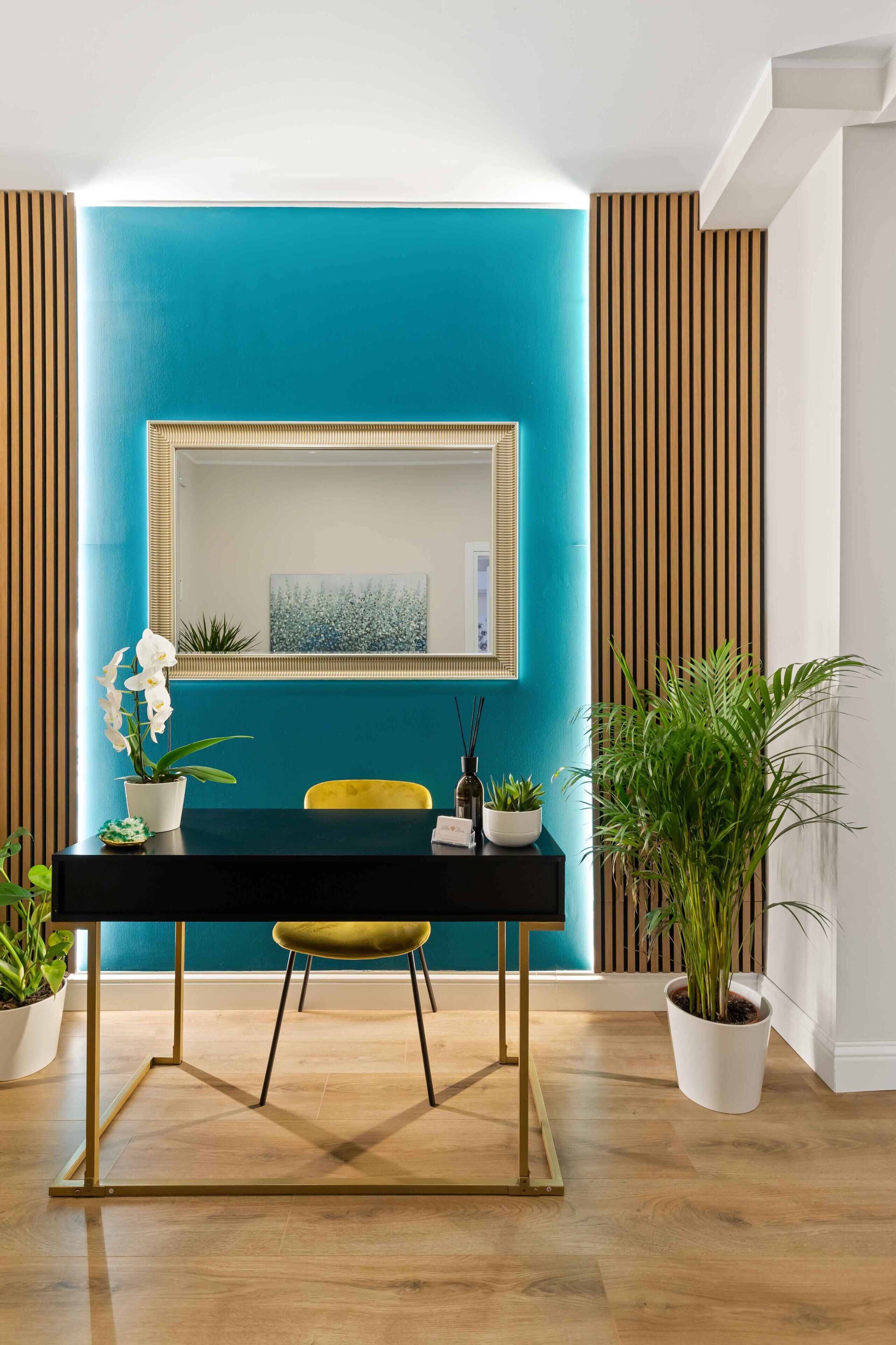 A modern room with a teal accent wall, a black desk with gold legs, a yellow chair, and potted plants.