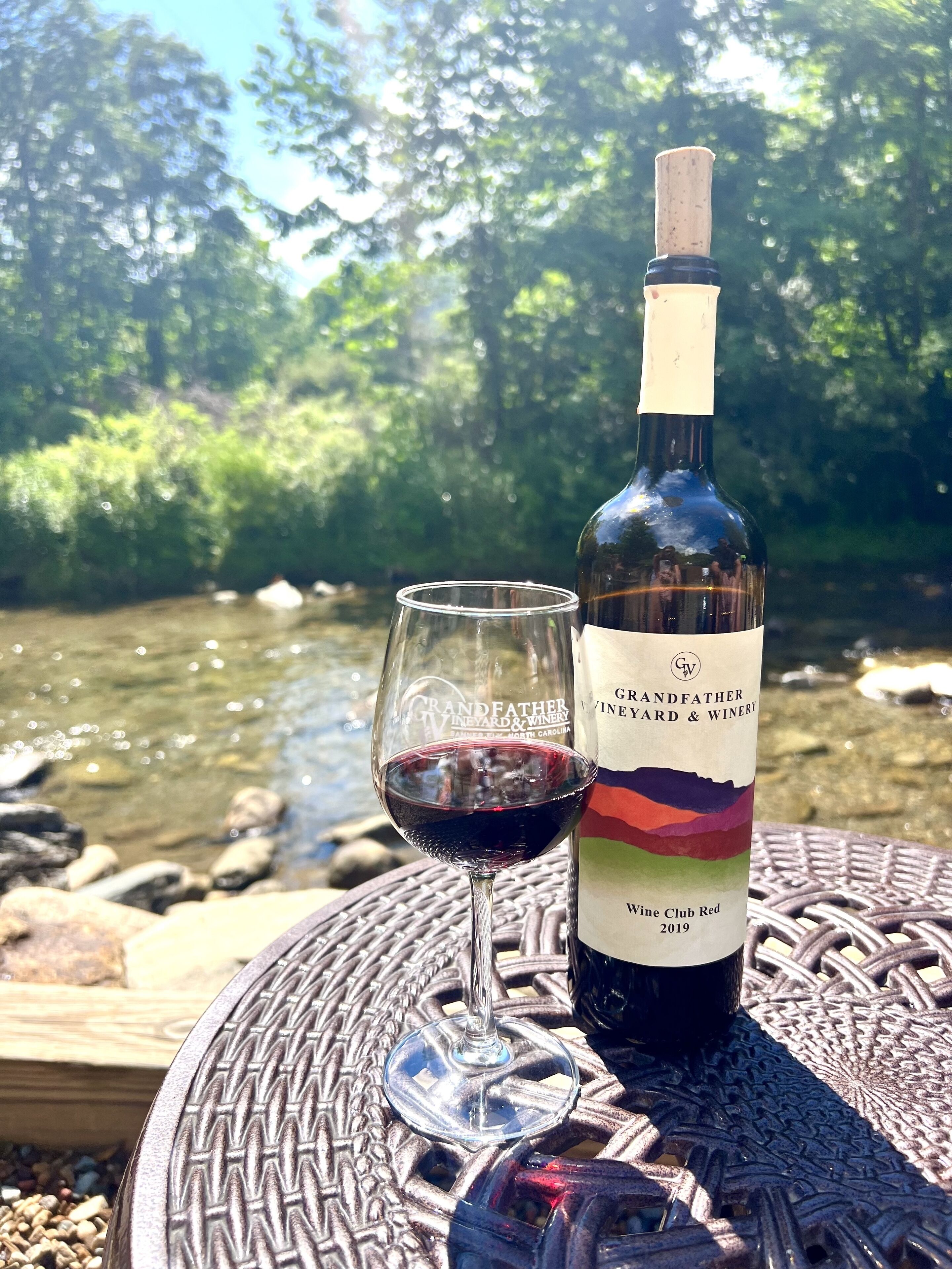 Una bottiglia di vino e un bicchiere su un tavolo con vista su un fiume e alberi sullo sfondo.