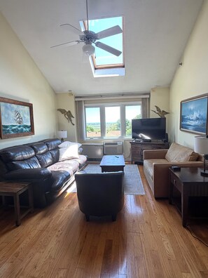 Un salon doté d’un ventilateur de plafond, d’une grande fenêtre, d’un canapé en cuir noir, d’une banquette en cuir marron, d’un téléviseur et d’un tableau représentant un navire accroché au mur.