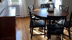 A dining area with a round wooden table, black chairs, and a chandelier. There are windows with curtains, a framed picture on the wall, and a vase on the table. - Beautiful Wells Maine Premium Top Floor Condo at Village By The Sea 2 Bed 2 Bath (Wells)