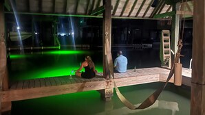 Een houten terras met een zwembad in groene verlichting, een hangmat en een persoon die aan de rand van het zwembad zit.