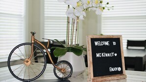 Una bicicleta, una maceta con flores blancas y una pizarra con la inscripción “WELCOME TO MCKINNEY” y “ENJOY”. 
