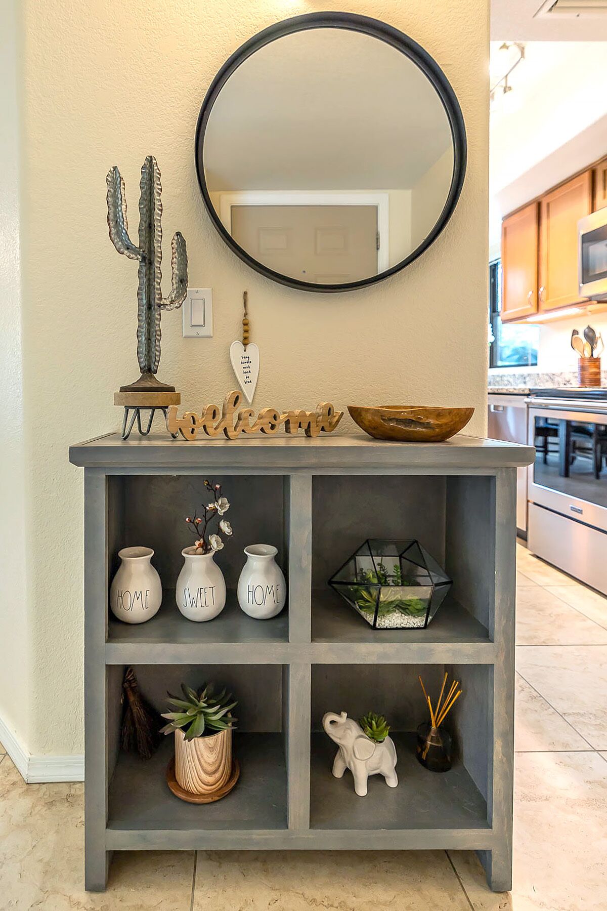 A grey console table with a mirror above it, decorated with a cactus, a heart-shaped sign, and a wooden welcome sign. Below, there are three white vases with 'HOME SWEET HOME' written on them, a plant in a wooden pot, and a small elephant figurine with a plant.