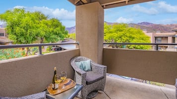 A balcony with a wicker chair, a table with a bottle and glasses, and a view of trees and mountains.