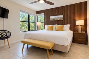 A bedroom with a large bed, two bedside lamps, a wooden headboard, a television, and a window with a view of trees. - Spacious Tucson Retreat • 3 King Beds • Pools/Spas, Mountain Views, Hiking/Golf (Tucson)