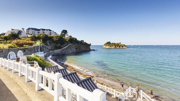 A coastal hotel with a beach promenade, a staircase leading to a cliff-top view, and a small island in the distance. - Les Bancs Roux - Binic Etables SUR MER (BINIC ETABLES SUR MER)