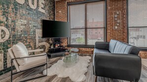 A living room with a brick wall, a flat-screen TV, a sofa, a chair, and a coffee table. - Deluxe Studio in Historic Downtown Building! (Bartlesville)