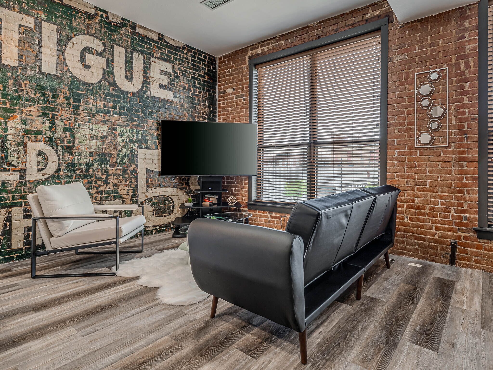 A living room with a brick wall, a large flat-screen TV, a black sofa, a white chair, and a wooden floor.