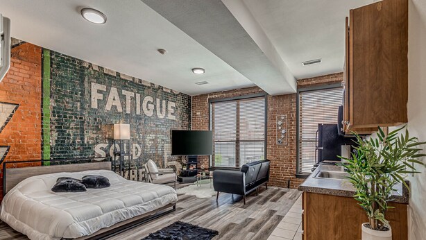 A modern bedroom with a large bed, a TV, a desk, and a brick wall with the word "FATIGUE" painted on it. - Deluxe Studio in Historic Downtown Building! (Bartlesville)