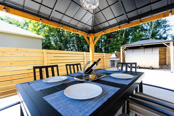 Un espacio de comedor al aire libre con una mesa negra, platos blancos y una botella de vino sobre un posavasos.