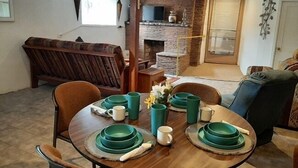 Uma área de refeições com uma mesa redonda de madeira posta com pratos verdes, xícaras e um vaso com flores. Há um banco de madeira ao fundo e uma lareira de tijolos.