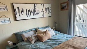 A bedroom with a bed, pillows, a nightstand, a wall sign, a window, and a view of a balcony. - The Posh Plum - 1 Block to the Casino Ballroom, Bars and Beach (Hampton)