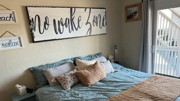Une chambre à coucher avec un lit, des oreillers, une table de chevet, un tableau décoratif au mur, une fenêtre et une vue sur un balcon.