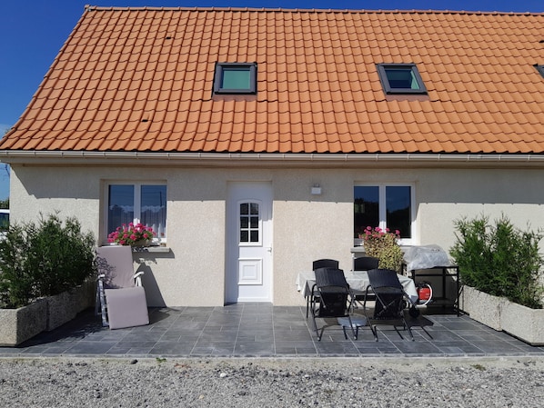 Une maison avec un toit en tuiles, une porte d’entrée, deux fenêtres, une terrasse avec des chaises et un barbecue, ainsi que des plantes en pot.
