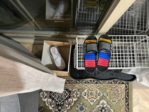 Sandals rest on a shelf above a traditional rug, with a view of a nearby building through the large window.
