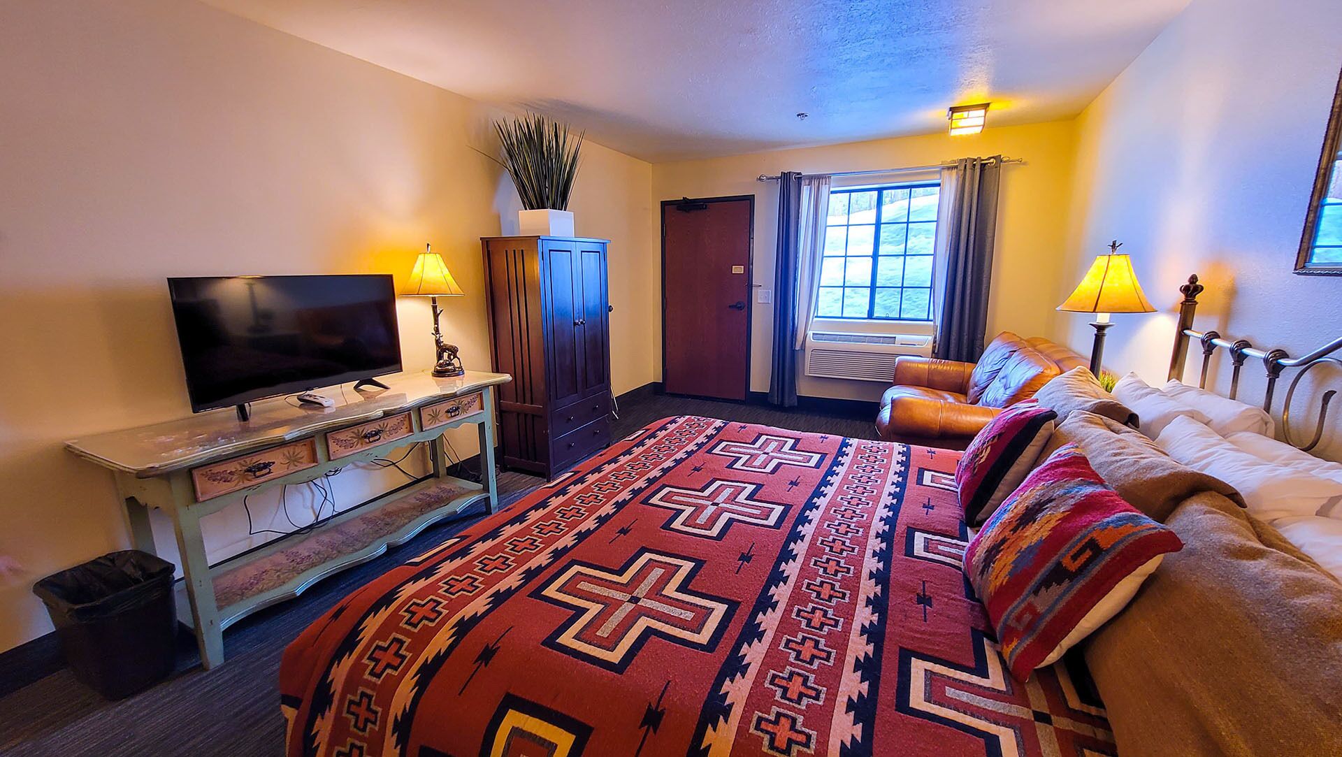 A hotel room with a large bed, a television, a desk with a lamp, and a window with curtains.