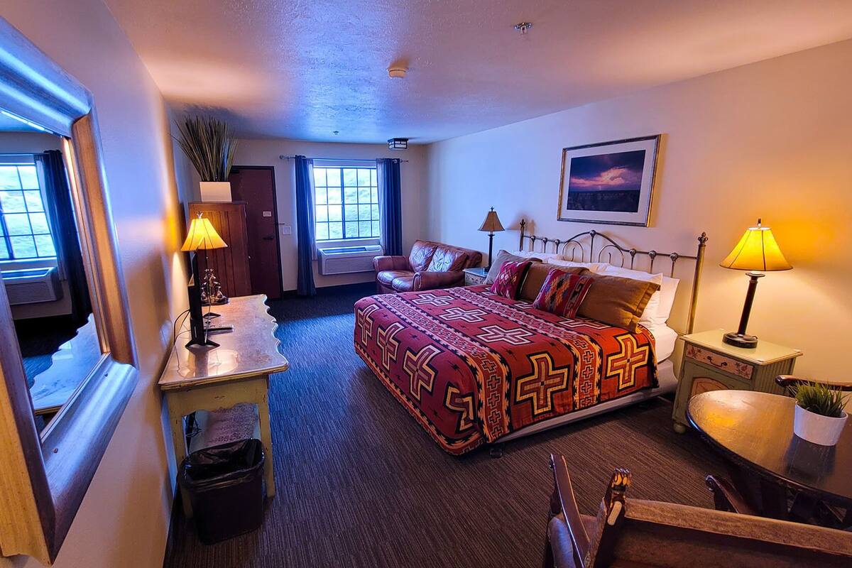 A hotel room with a large bed, two bedside lamps, a window with curtains, a small table with a lamp, and a framed picture on the wall.