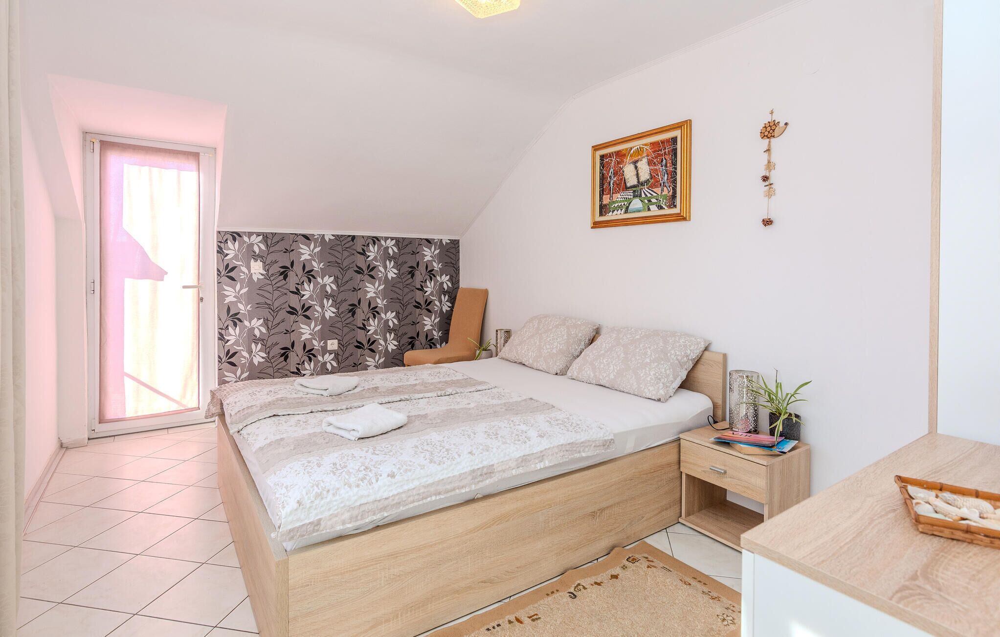 Een slaapkamer met een bed, een nachtkastje, een stoel, een raam en een schilderij aan de muur.