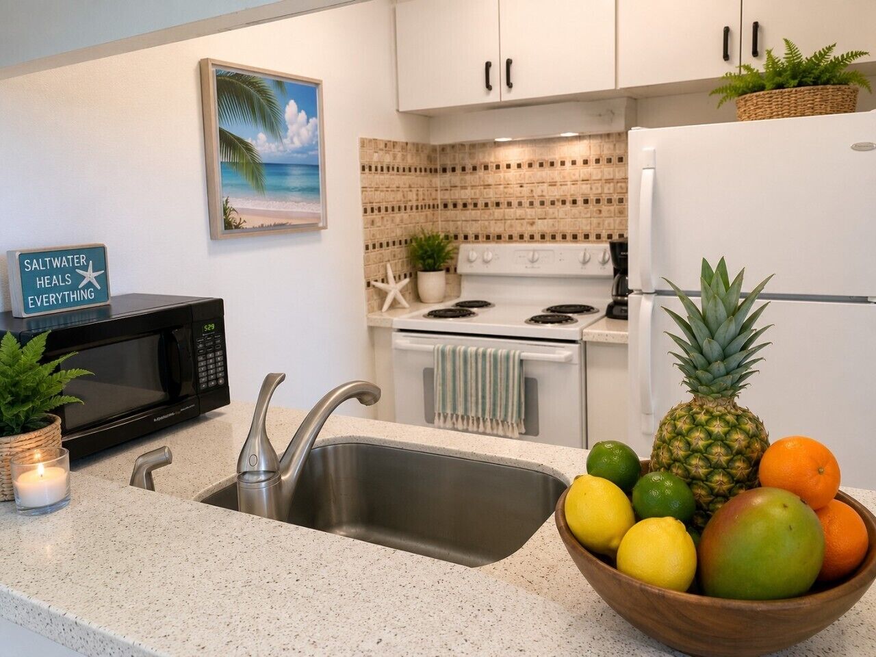 Una cocina con encimera de granito, pileta de acero inoxidable, muebles blancos, microondas, estufa y un tazón de fruta.