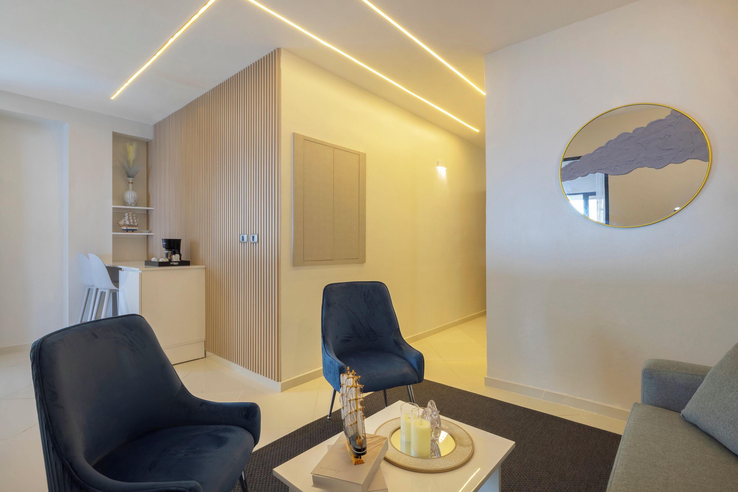 Ein modernes Wohnzimmer mit zwei dunkelblauen Sesseln, einem weißen Couchtisch mit Dekoration, einem runden Spiegel und einem hellen Sofa.