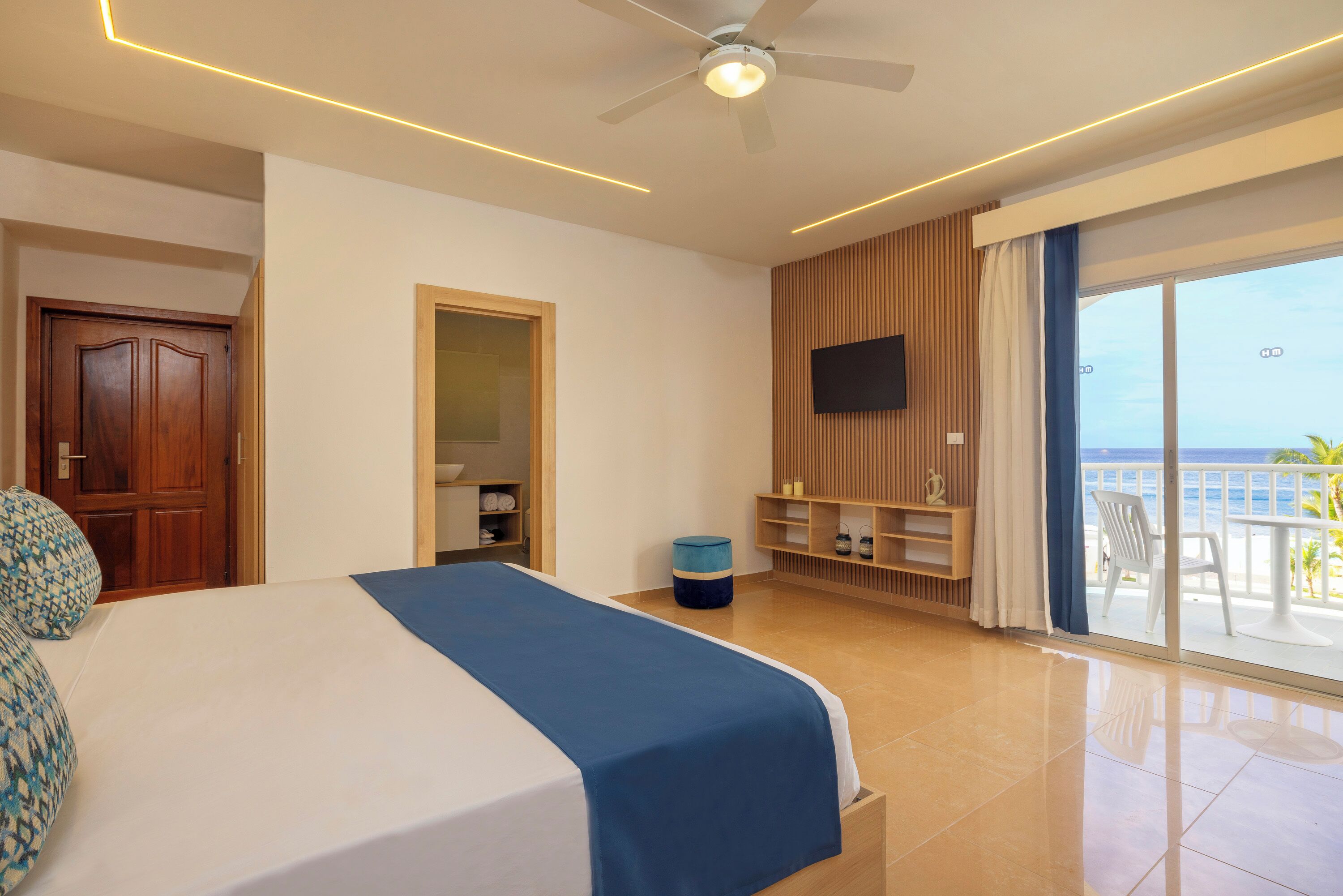 A hotel room with a bed, a TV mounted on the wall, a balcony with a view of the sea, and a ceiling fan.